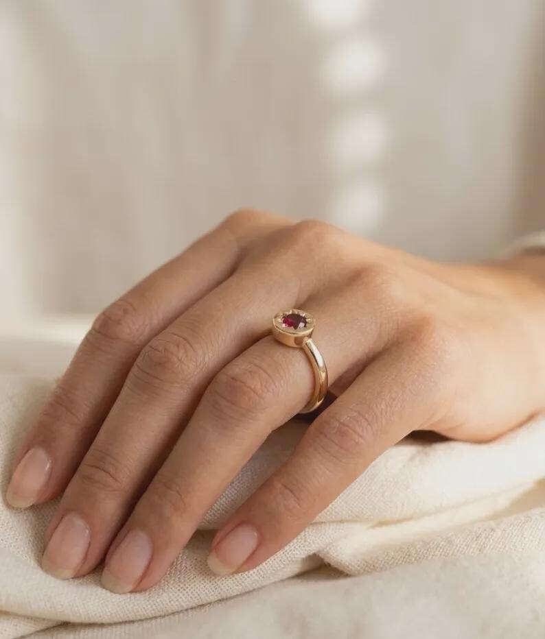 May include: A gold ring featuring a round, deep red gemstone set in a circular bezel. The band is smooth and polished, showcasing a simple, clean design that highlights the vibrant color of the central stone and the warm tone of the metal.