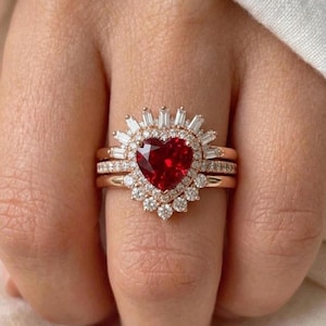 May include: A rose gold ring set with a heart-shaped, deep red gemstone at its centre, encircled by small diamonds. The main ring is complemented by two additional bands, creating a layered appearance. The ring's design is elegant and striking.