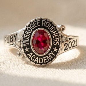 以下が含まれることがあります： 中央にオーバルカットの赤い石を配し、MUSKOGEE ROUGHERS ACADEMYの文字が刻まれた黒い縁取りが施されたシルバーのクラスリング。バンドの片側にはRUBY、もう片側には2021と刻印されており、2つの小さな透明な石がアクセントになっています。