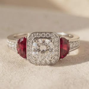 Peut inclure: Une bague argentée avec une pierre centrale transparente taille coussin entourée d'un halo de petites pierres transparentes rondes. Deux pierres rouges en demi-lune sont placées de chaque côté, avec des pierres transparentes sur l'anneau.