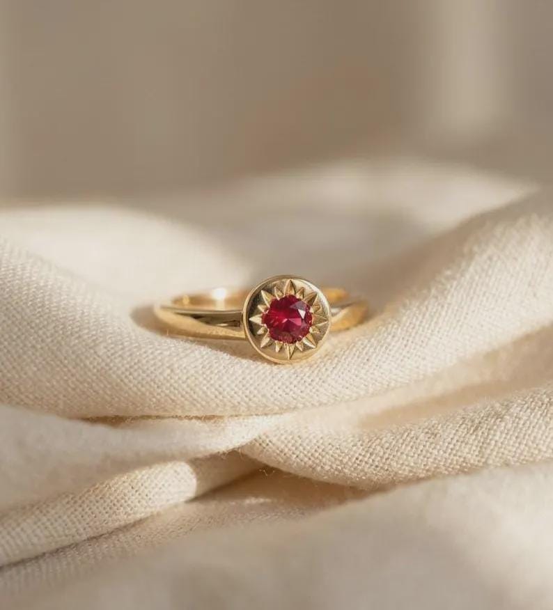 Gold ring featuring a central round red ruby set in an engraved sunburst or star-style design on a textured fabric background.