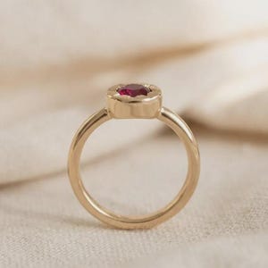 May include: A gold ring featuring a round, faceted deep red gemstone set in a circular bezel setting. The band is smooth and polished, with the gemstone positioned at the top of the ring, showcasing a vibrant red color and a metallic gold finish.