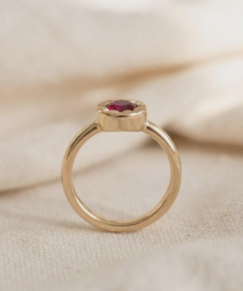 May include: A gold ring featuring a round, faceted deep red gemstone set in a circular bezel setting. The band is smooth and polished, with the gemstone positioned at the top of the ring, showcasing a vibrant red color and a metallic gold finish.