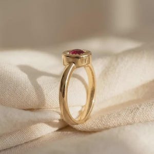 May include: A gold ring featuring a smooth, polished band and a circular setting. The center of the setting holds a small, round, faceted red gemstone. The ring is positioned upright, showcasing the profile of the band and the gemstone setting.