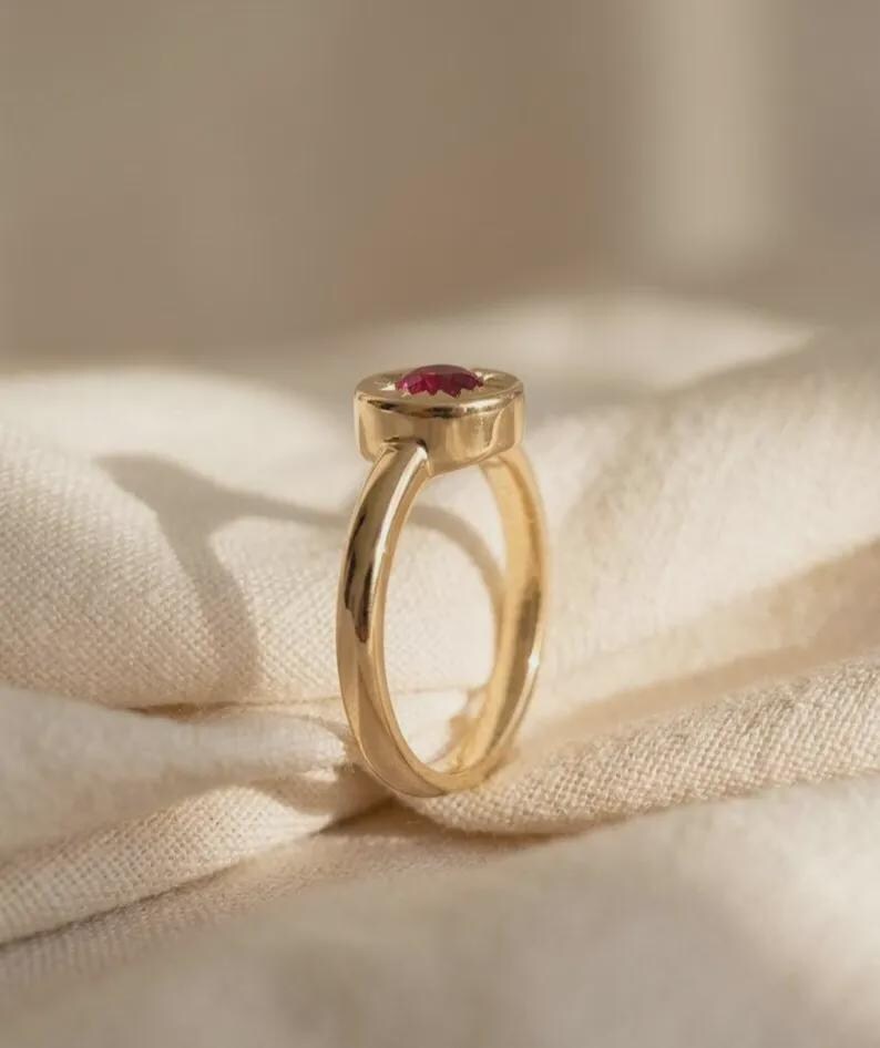 May include: A gold ring featuring a smooth, polished band and a circular setting. The center of the setting holds a small, round, faceted red gemstone. The ring is positioned upright, showcasing the profile of the band and the gemstone setting.