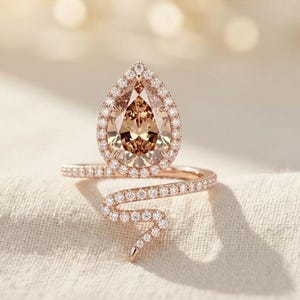 May include: A rose gold ring featuring a pear-shaped champagne-coloured centre stone surrounded by a halo of small white stones. The band is designed in a serpentine shape, fully encrusted with tiny white stones that extend along the entire length.