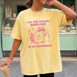 May include: A pale yellow short-sleeved t-shirt featuring a pink graphic of a raccoon wearing sunglasses, holding a martini glass, and carrying a baguette. The text reads I AM THE PRIMARY BREADLOSER OF MY HOUSEHOLD in pink capital letters.