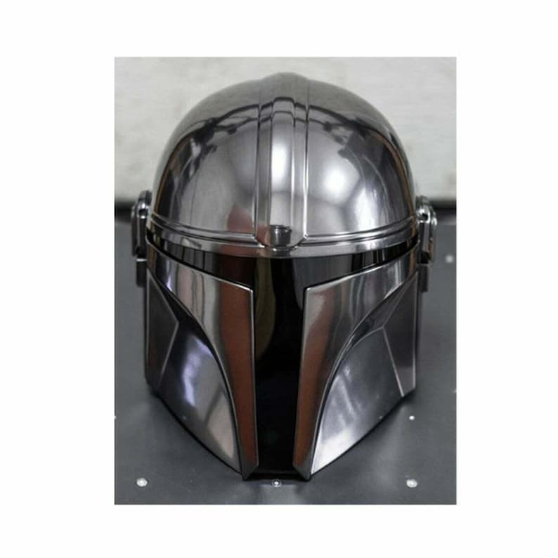 May include: A metallic silver Mandalorian helmet with a black visor. The helmet has a smooth, reflective surface with vertical ridges on the top. The design features angular shapes and a distinctive T-shaped visor.