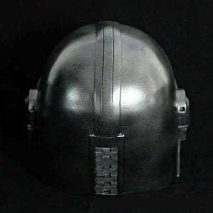 May include: A metallic silver helmet with a rounded top and vertical ridges. The helmet has side details and a rectangular section at the back with a series of small, square shapes. The overall design suggests a futuristic or industrial aesthetic.
