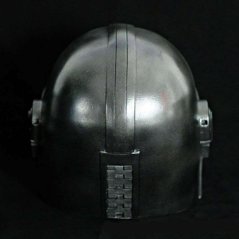 May include: A metallic silver helmet with a rounded top and vertical ridges. The helmet has side details and a rectangular section at the back with a series of small, square shapes. The overall design suggests a futuristic or industrial aesthetic.