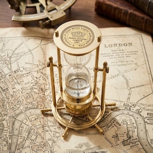 Puede incluir: Un reloj de arena de latón con una base en forma de timón de barco y una placa superior grabada con "A journey of 1000 Miles begins with a single step" y "HENRY HUGHES LONDON 1941". El reloj tiene bulbos de vidrio transparente y marco metálico.