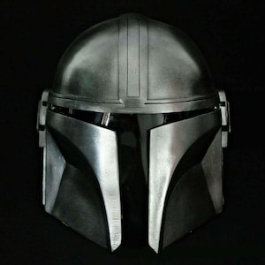 May include: A metallic silver Mandalorian helmet with a dark visor. The helmet has a rounded top with vertical ridges and angular cheek plates. The helmet is designed with a sleek, futuristic aesthetic, perfect for cosplay or display.
