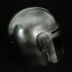 May include: A metallic silver helmet with a visor and side details. The helmet has a slightly weathered appearance, suggesting a battle-worn aesthetic. The design is reminiscent of a sci-fi or space-themed character.