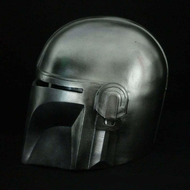 May include: A metallic silver Mandalorian helmet, featuring a T-visor and distinctive cheek and jaw design. The helmet has a smooth, reflective surface and is set against a dark background. The helmet is a replica of the one worn by the character in the Star Wars series.