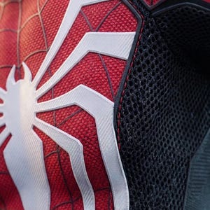 May include: Close-up of a red and black Spider-Man costume. The costume features a white spider emblem on the chest and white web patterns. The suit has a textured fabric and black mesh detailing.