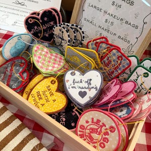 May include: A wooden crate filled with assorted embroidered fabric tags and bookmarks in various shapes, including hearts and circles. The items feature colourful patterns, floral prints, and text such as "fuck off i'm reading" and "SAVE A HORSE RIDE A COWBOY."