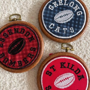May include: Three circular wooden embroidery hoops featuring fabric inserts with embroidered Australian football team logos. One hoop displays Geelong Cats, one Essendon Bombers, and one St Kilda Saints, each with a central football graphic.