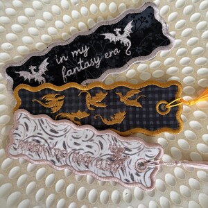 May include: Three rectangular fabric bookmarks with scalloped edges. The top one is black with white dragon silhouettes and the text 'in my fantasy era'. The middle one is dark grey plaid with gold dragon embroidery. The bottom one is white with grey swirls.