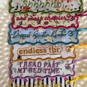 May include: A set of six rectangular fabric bookmarks with scalloped edges and embroidered text. The designs feature floral patterns, stripes, and phrases including Shhhhh..., one more chapter, Dearest Gentle Reader, endless tbr, READ PAST MY BED TIME, and The Book Lovers Club.
