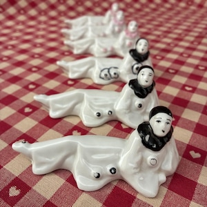 May include: A collection of white porcelain figurines, each depicting a person in a reclining pose. The figures have black accents on their clothing and hair, and are arranged on a red and white checkered surface.