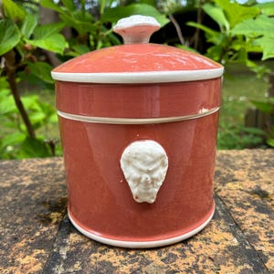 May include: A terracotta-colored ceramic jar with a lid and a white decorative accent. The jar has a white rim and a white relief design on the front. The lid has a white knob. The jar is cylindrical and sits on a rough surface.