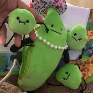 May include: A whimsical plush pea pod with a zipper, adorned with a pearl necklace, surrounded by three plush peas. The peas and pod are light green with black facial features and black string arms and legs. A floral patterned item is in the background.