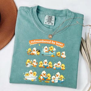 Peut inclure: Un t-shirt Comfort Colors bleu sarcelle avec l'inscription "outnumbered by boys" en orange. Le t-shirt présente un graphisme de canards de dessins animés dans diverses scènes. Un collier argenté avec un pendentif est drapé sur le t-shirt.
