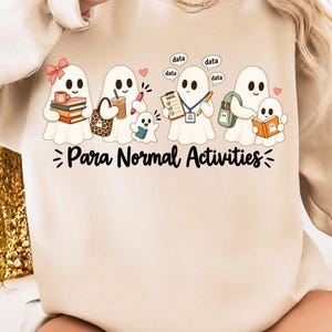 May include: A beige crewneck sweatshirt featuring a graphic of five white ghosts engaged in various tasks. The text Para Normal Activities is printed below the ghosts. One ghost holds books, another a coffee cup, a clipboard, and a backpack.