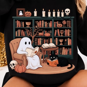 Könnte beinhalten: Ein schwarzes Sweatshirt mit einer grafischen Illustration eines weißen Geistes, der in einem braunen Sessel sitzt und ein Buch liest, einer schwarzen Katze, die auf einem Teppich liest, einem Bücherregal voller Bücher, Kerzen, Kürbissen und einem Totenkopf.