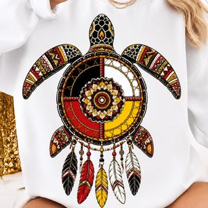 May include: A white sweatshirt featuring a large graphic of a sea turtle with a shell designed as a dreamcatcher. The turtle has patterned flippers in red, black, and yellow. Five feathers in black, red, yellow, and white hang from the bottom.