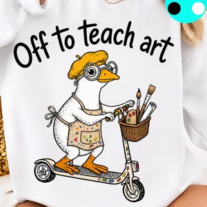 May include: A white sweatshirt featuring a graphic of a white goose wearing a yellow beret, round glasses, and a paint-splattered apron. The goose is riding a scooter with a basket containing paintbrushes and a palette. The text reads Off to teach art.