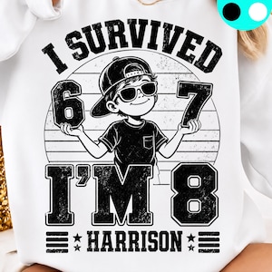 Könnte beinhalten: Ein weißes Sweatshirt mit einem schwarzen Grafikdruck eines Kindes mit Baseballkappe und Sonnenbrille, das die Zahlen 6 und 7 hält. Der Text lautet I SURVIVED, I'M 8 und HARRISON, ergänzt durch ein kreisförmiges Design mit Used-Optik.