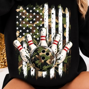 May include: A black long-sleeve sweatshirt featuring a graphic print of a bowling ball and four bowling pins arranged in front of a distressed American flag design with a green, brown, and black camouflage pattern and white stars and stripes.