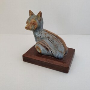 May include: A ceramic fox figurine in shades of blue, brown, and tan, mounted on a rectangular wooden base. The fox is in a seated position, with its ears perked up. The base is approximately 4 inches long.