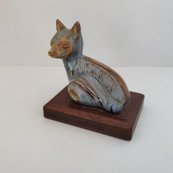 Może przedstawiać: Ceramiczna figurka lisa w odcieniach niebieskiego, brązowego i beżowego, osadzona na prostokątnej drewnianej podstawie. Lis siedzi, z uniesionymi uszami. Podstawa ma około 10 cm długości.