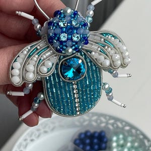 Op de afbeelding: Handgemaakte insectenbroche van kralen in blauwe, turquoise en witte tinten. De kevervormige speld heeft een grote, gefacetteerde blauwe edelsteen en ingewikkeld kralenwerk, met zilveren draaddetails. De vleugels zijn versierd met witte kralen.