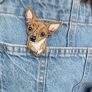 Pode incluir: Broche de Chihuahua artesanal, bordado com fios castanhos, dourados e brancos. A cabeça do cão é detalhada com olhos pretos e um nariz preto, acentuado com pérolas rosa nas orelhas. O broche está preso a um bolso de ganga azul claro.