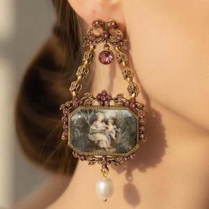 May include: A gold-toned drop earring featuring an ornate metal frame with small pink crystals, a central rectangular plaque depicting a classical scene, and a single white pearl dangling from the bottom of the rectangular frame.