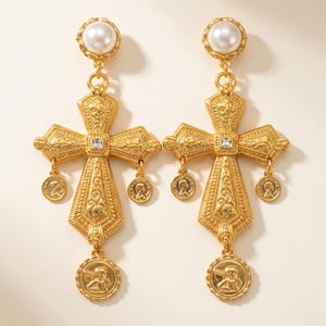 May include: A pair of gold-toned drop earrings featuring a large ornate cross pendant with intricate scrollwork patterns. Each earring is topped with a round white pearl and includes three small circular coin charms dangling from the cross arms.