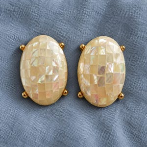 May include: A pair of oval-shaped earrings featuring a mosaic pattern of iridescent mother-of-pearl tiles. Each earring is framed by a polished gold-tone metal border with four small, rounded gold-tone accents positioned at the cardinal points.