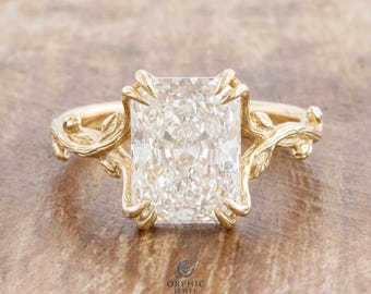 Op de afbeelding: Een gouden ring met een grote, rechthoekige, heldere edelsteen in radiant-slijpvorm, vastgehouden door acht pootjes. De band is ontworpen als verstrengelde boomtakken met kleine, gebeeldhouwde blaadjes langs de zijkanten van de zetting.