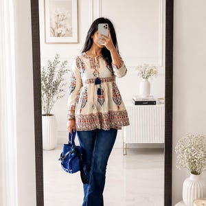 Puede incluir: Una túnica color crema con intrincados bordados florales en rojo, azul y verde, combinada con unos vaqueros acampanados de lavado oscuro. Un bolso cubo azul y sandalias doradas completan el conjunto. La imagen se refleja en un espejo grande.