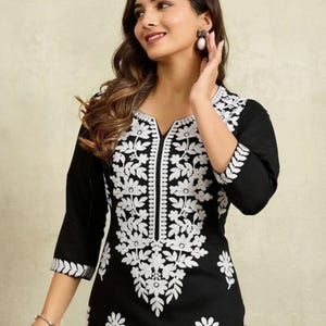 Könnte beinhalten: Schwarze Tunika mit weißer Blumenstickerei. Die Tunika hat einen V-Ausschnitt und Dreiviertelärmel. Die weiße Stickerei ist detailliert und bedeckt den Ausschnitt, die Vorderseite und die Ärmel. Das Kleidungsstück ist aus weichem Stoff.