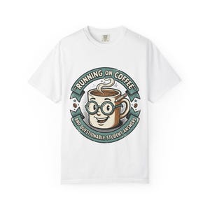 Puede incluir: Camiseta blanca con un diseño de taza de café de dibujos animados. La taza tiene gafas y un lápiz, con el texto "RUNNING ON COFFEE AND QUESTIONABLE STUDENT ANSWERS" en un diseño circular. La taza es marrón.