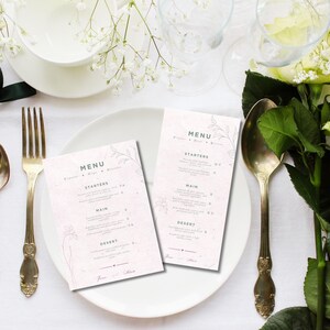 Peut inclure: Mise en place pour un mariage avec une assiette blanche, des couverts dorés et deux menus. Les menus présentent un motif floral et le mot « MENU ». Des fleurs blanches et du feuillage vert ornent la table.
