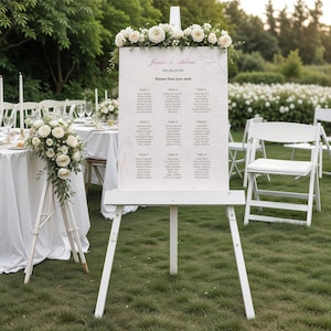 Peut inclure: Un chevalet blanc affichant un plan de table de mariage, orné de compositions florales. Le plan comporte des noms et des affectations de tables, avec les noms "Janie & Adam" et la date "06.26.2020" en haut. La scène se déroule en extérieur avec des chaises et des tables blanches.