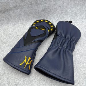 May include: Two navy blue golf club head covers. One cover features a black and navy blue design of a hooded figure with yellow star accents and a yellow monogram. The other cover is navy blue.