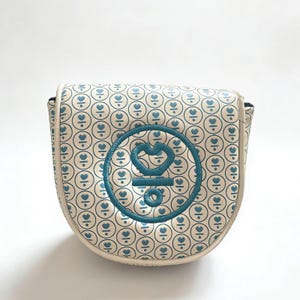 May include: Cream-colored golf club head cover with a repeating pattern of blue hearts inside circles. A larger blue embroidered emblem is centered on the cover. The cover has a rounded shape and a white border.