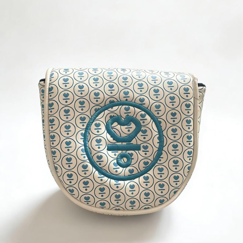 May include: Cream-colored golf club head cover with a repeating pattern of blue hearts inside circles. A larger blue embroidered emblem is centered on the cover. The cover has a rounded shape and a white border.
