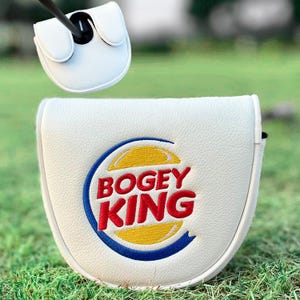 Może przedstawiać: Biała osłona gł&oacute;wki kija golfowego z napisem "BOGEY KING" w kolorze czerwonym, ż&oacute;łtym i niebieskim. Osłona znajduje się na zielonej, trawiatej powierzchni. Kij golfowy jest włożony w osłonę.